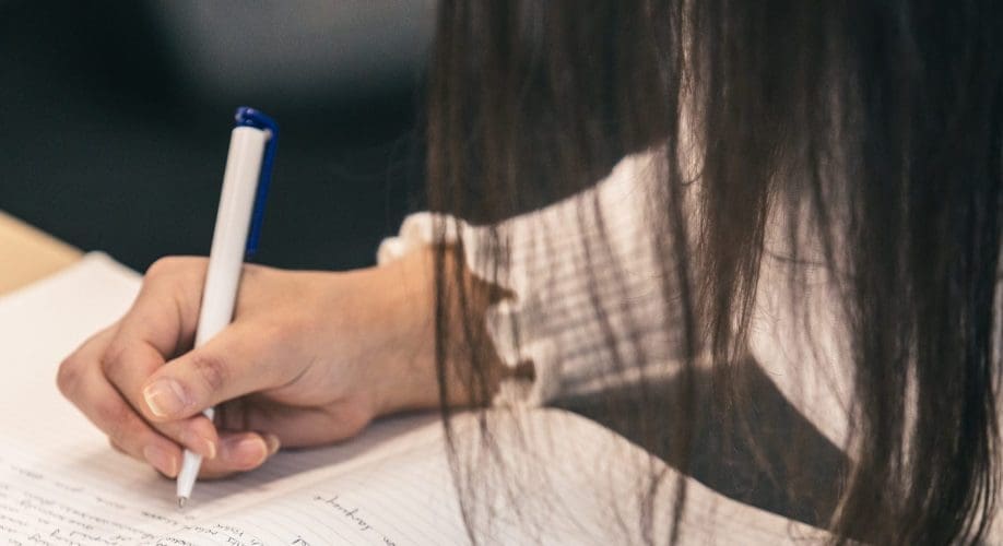 A person with long dark hair writes in a notebook with a blue and white pen. The photo focuses on their hand and the page filled with handwritten notes.